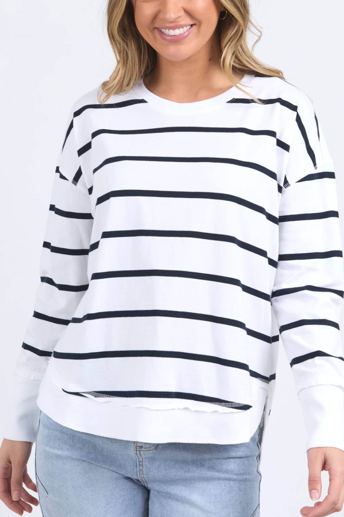 A woman wears a Foxwood Farrah Stripe Long Sleeve in white with black horizontal stripes and light blue jeans, smiling against a plain background.