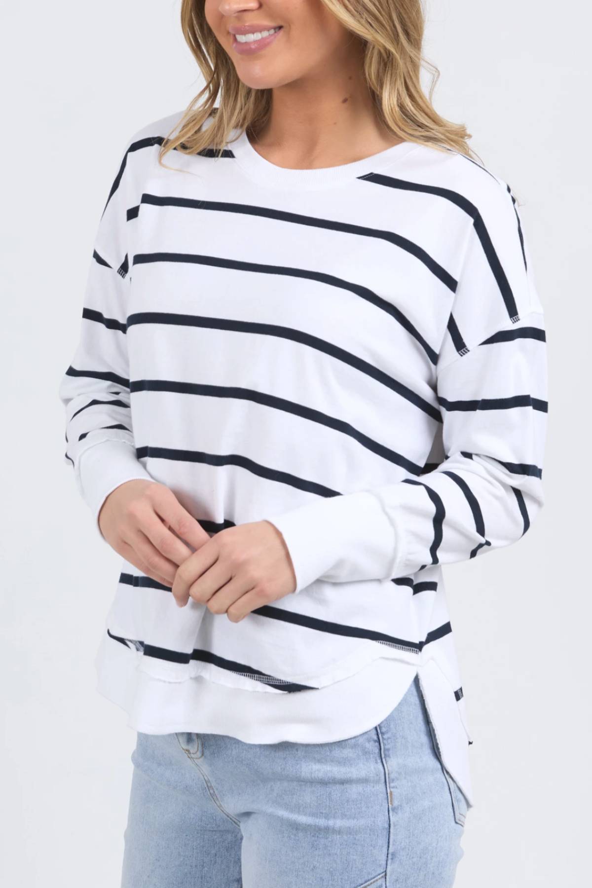 A woman wears the Foxwood Farrah Stripe Long Sleeve with black stripes and light blue jeans. She stands smiling, hands together, with loose, wavy hair.