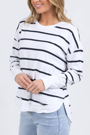 A woman wears the Foxwood Farrah Stripe Long Sleeve with black stripes and light blue jeans. She stands smiling, hands together, with loose, wavy hair.