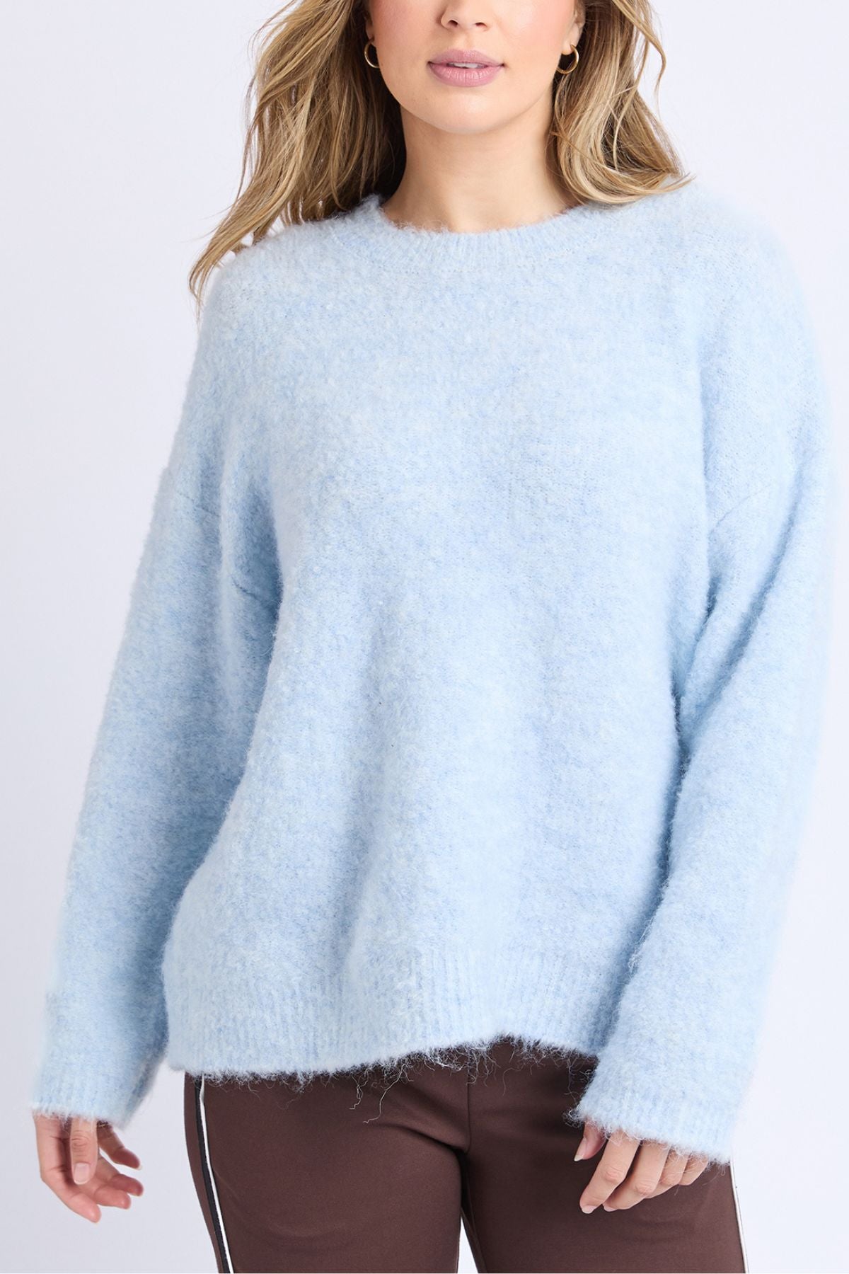 A woman in a soft, light blue Foxwood Flint Knit Crew sweater and brown pants with white side stripes stands against a plain white background, her hair loose and body slightly turned to the side.