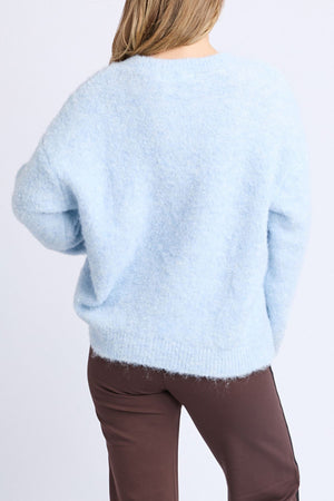 A person with light brown hair is wearing the Foxwood Flint Knit Crew sweater and brown pants, facing away from the camera against a plain light background.