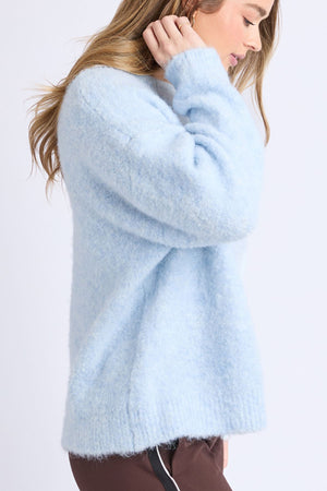A person with long, light brown hair wears the Foxwood Flint Knit Crew sweater in soft, light blue and brown pants. They stand sideways, touching their ear and looking slightly downward.