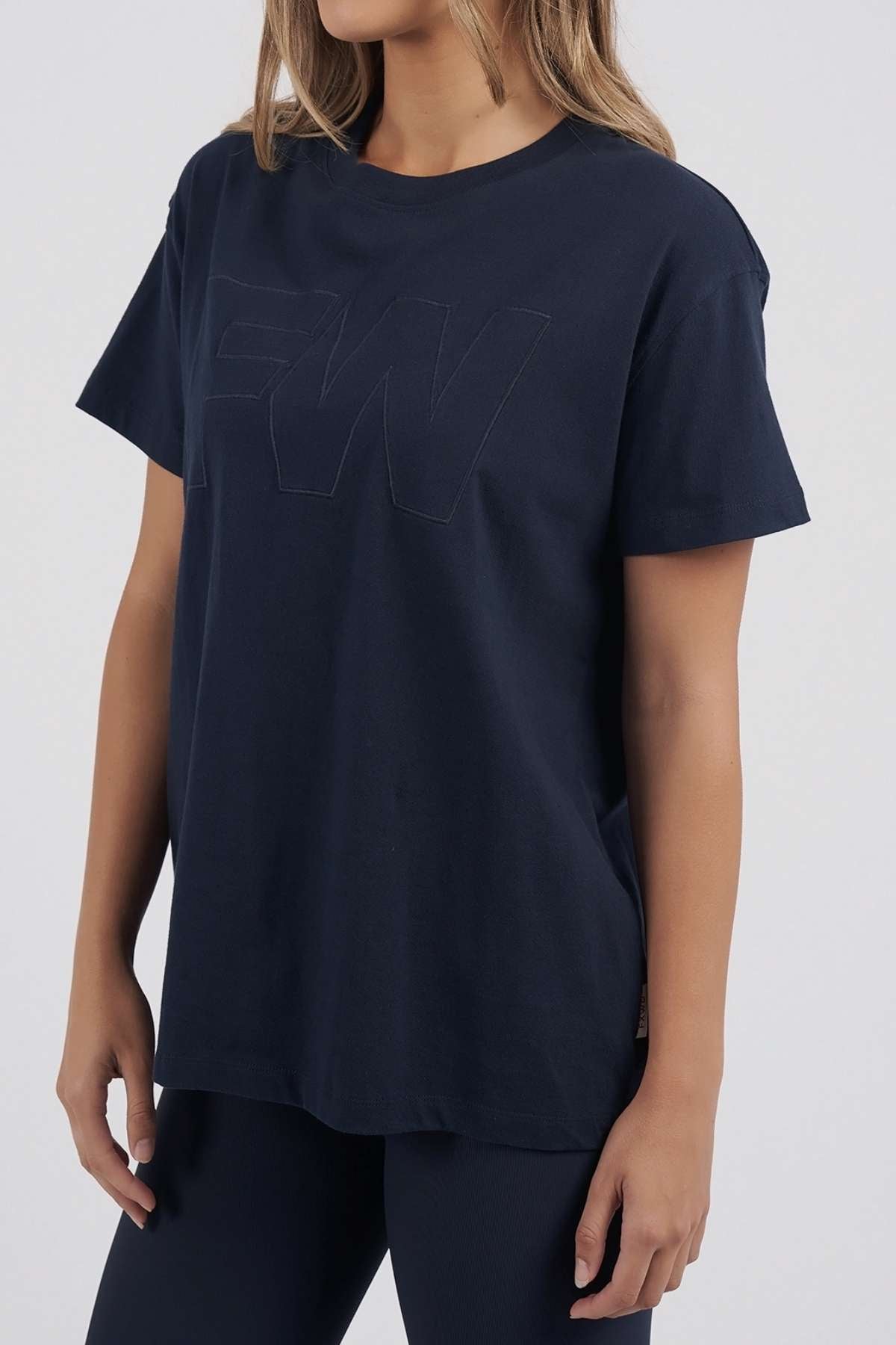 A woman wears the navy Foxwood FW Embroidery Tee, a loose-fitting 100% cotton top with short sleeves and subtle satin "FW" embroidery on the chest, paired with dark pants. Her face is partially out of frame.