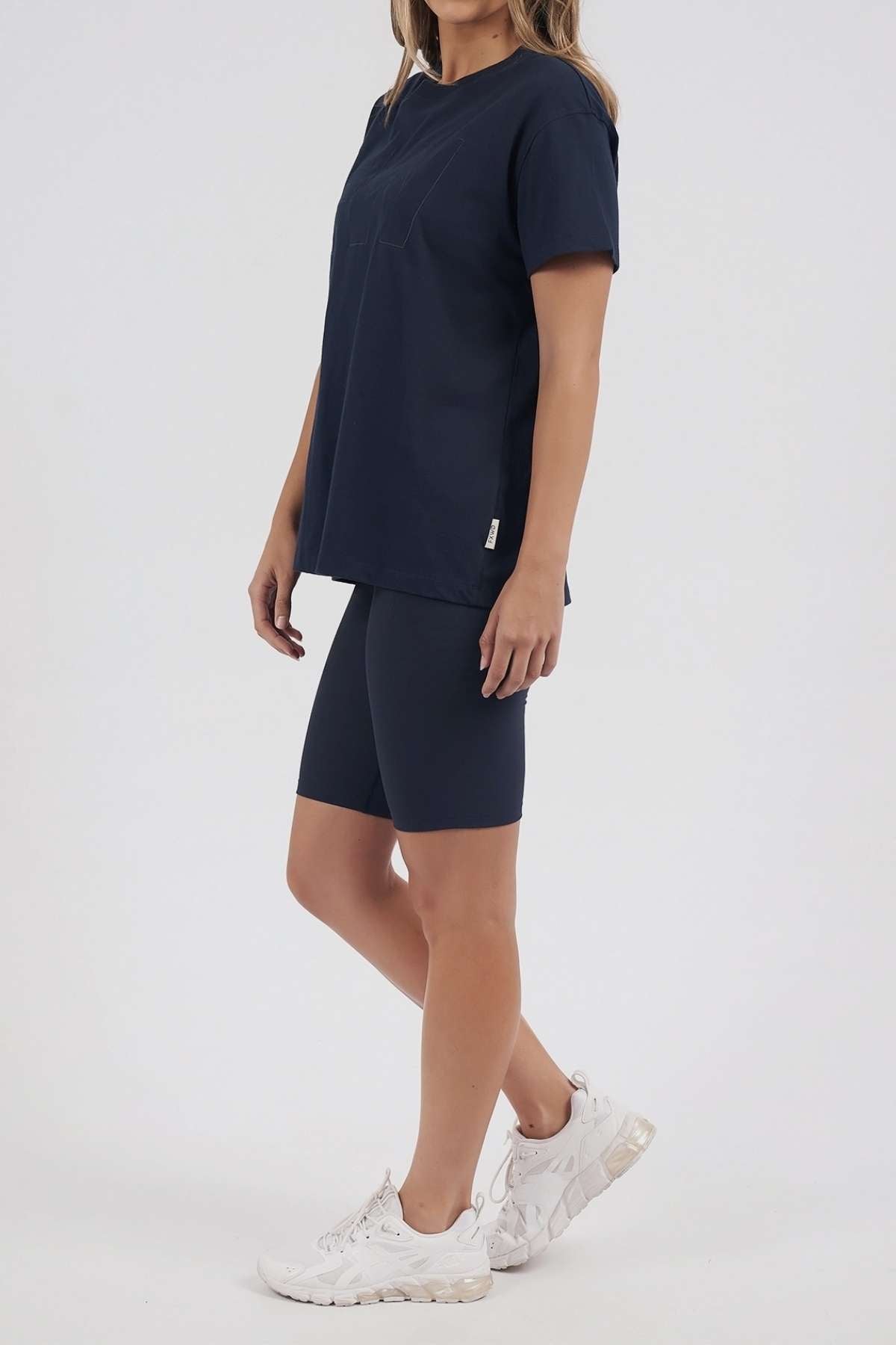 A person stands against a plain light background, wearing a navy blue Foxwood FW Embroidery Tee with satin embroidery, matching navy blue bike shorts, and white sneakers. The tee is crafted from 100% cotton. The face is not visible.