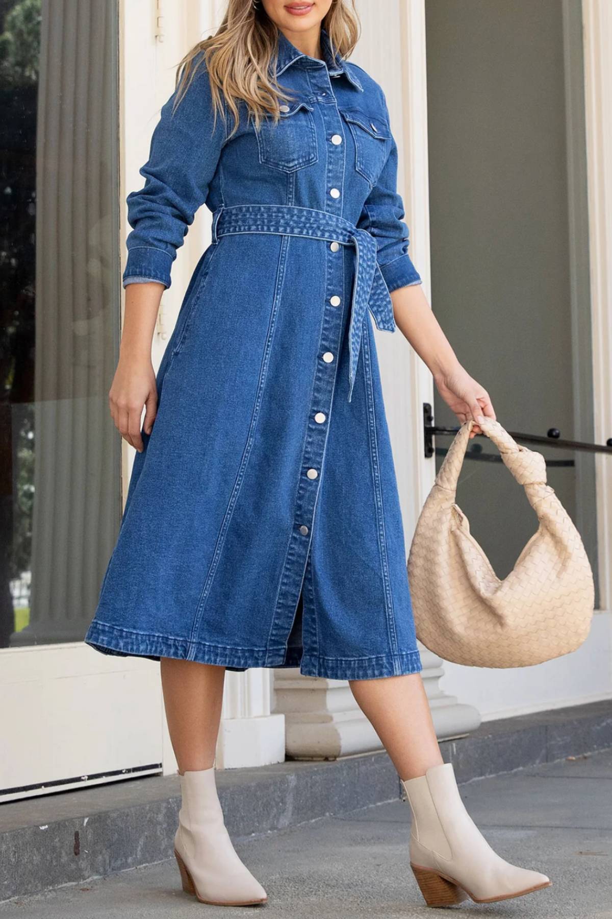 A woman wears the Foxwood Genevieve Denim Dress with a matching belt, paired with a large woven beige handbag and light beige ankle boots, standing outdoors in front of a building for an effortlessly stylish look.