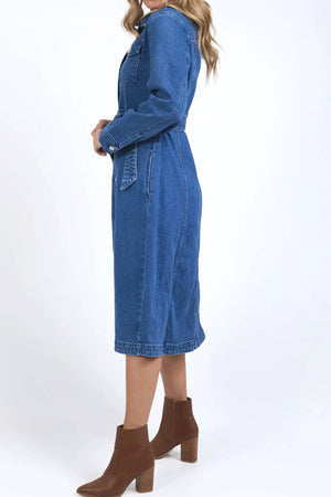A woman stands sideways in the Foxwood Genevieve Denim Dress—midi length with long sleeves and a belted waist—paired with brown ankle boots against a plain white background.