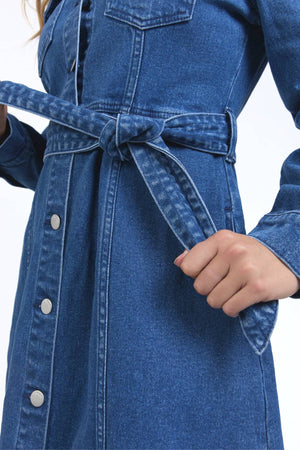 A person ties the waist belt of the Foxwood Genevieve Denim Dress, a blue midi dress with metal buttons and long sleeves, showcasing its stylish and functional design. The image highlights the hands and torso.