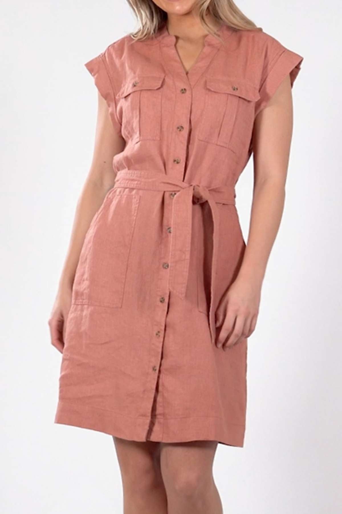 The individual is in a pink Foxwood Harlow Dress, featuring short sleeves, knee-length design, a button-up front with a fabric belt, and two chest pockets. Its flattering silhouette beautifully contrasts against the plain light backdrop.