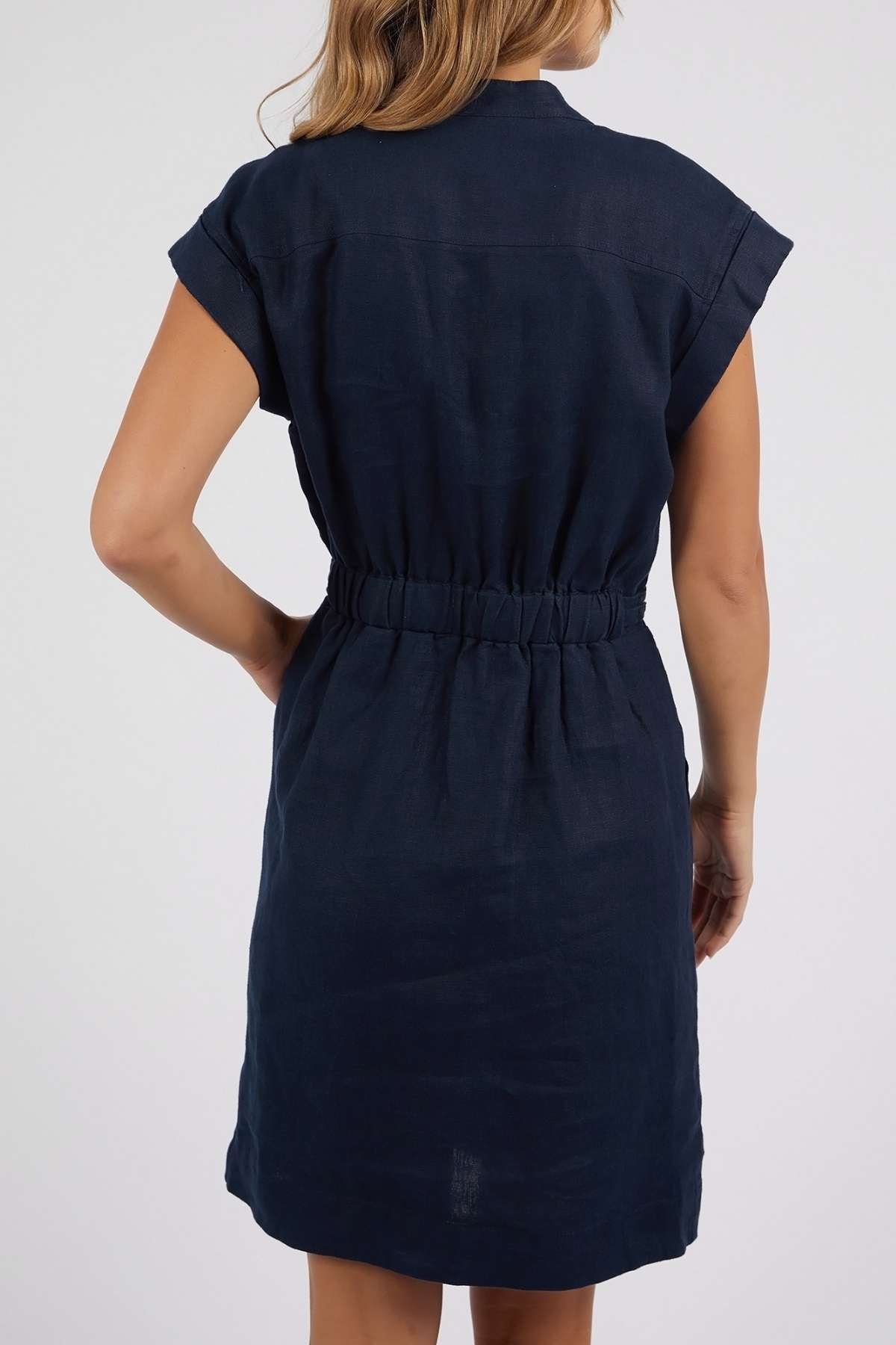 A woman with light brown hair is seen from the back wearing the Foxwood Harlow Dress, a short-sleeved, knee-length navy piece with a v-neckline and elastic waistband, set against a plain light background.