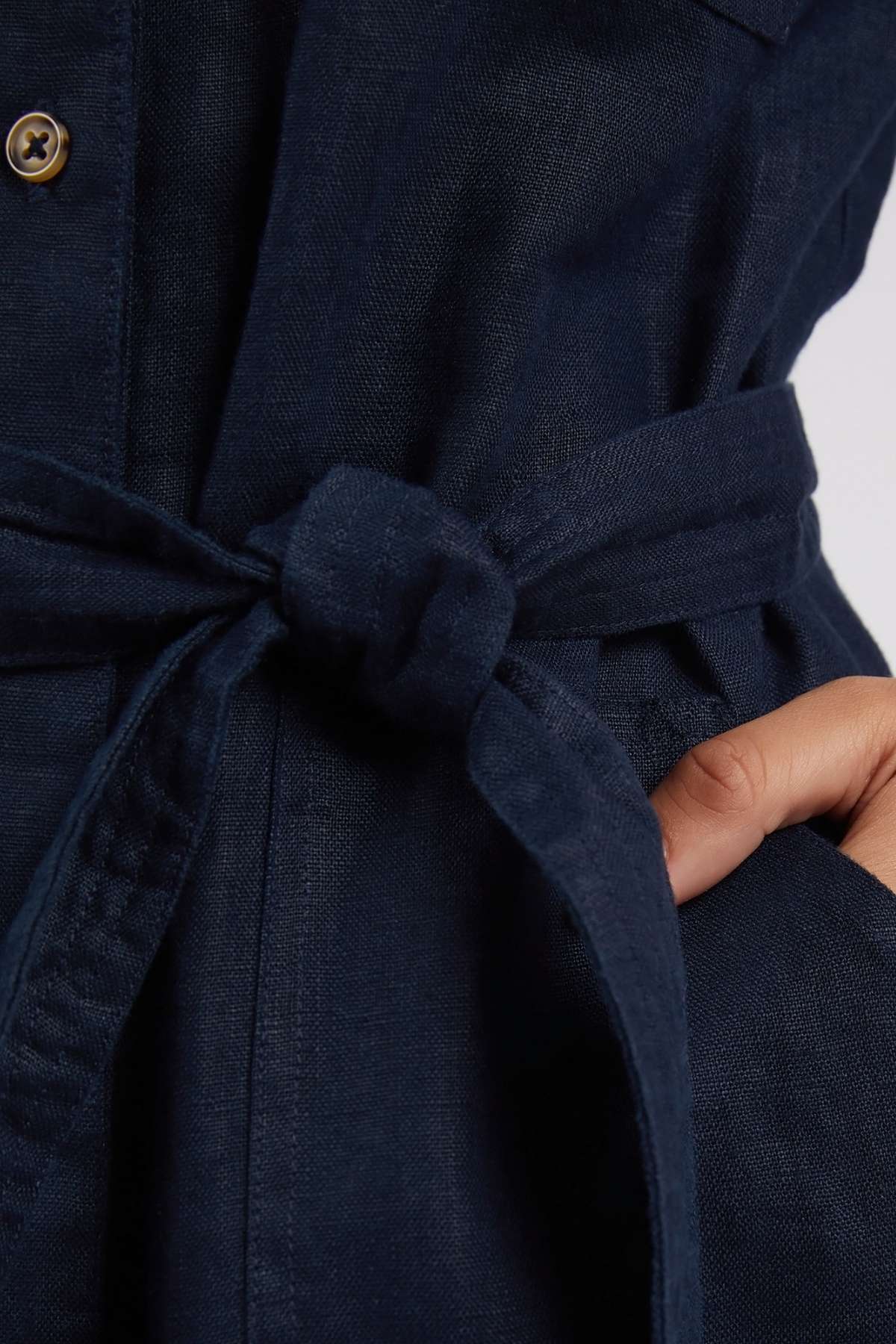 A close-up of a hand in the pocket of a Foxwood Harlow Dress, featuring a dark blue color and a fabric belt knotted at the waist.