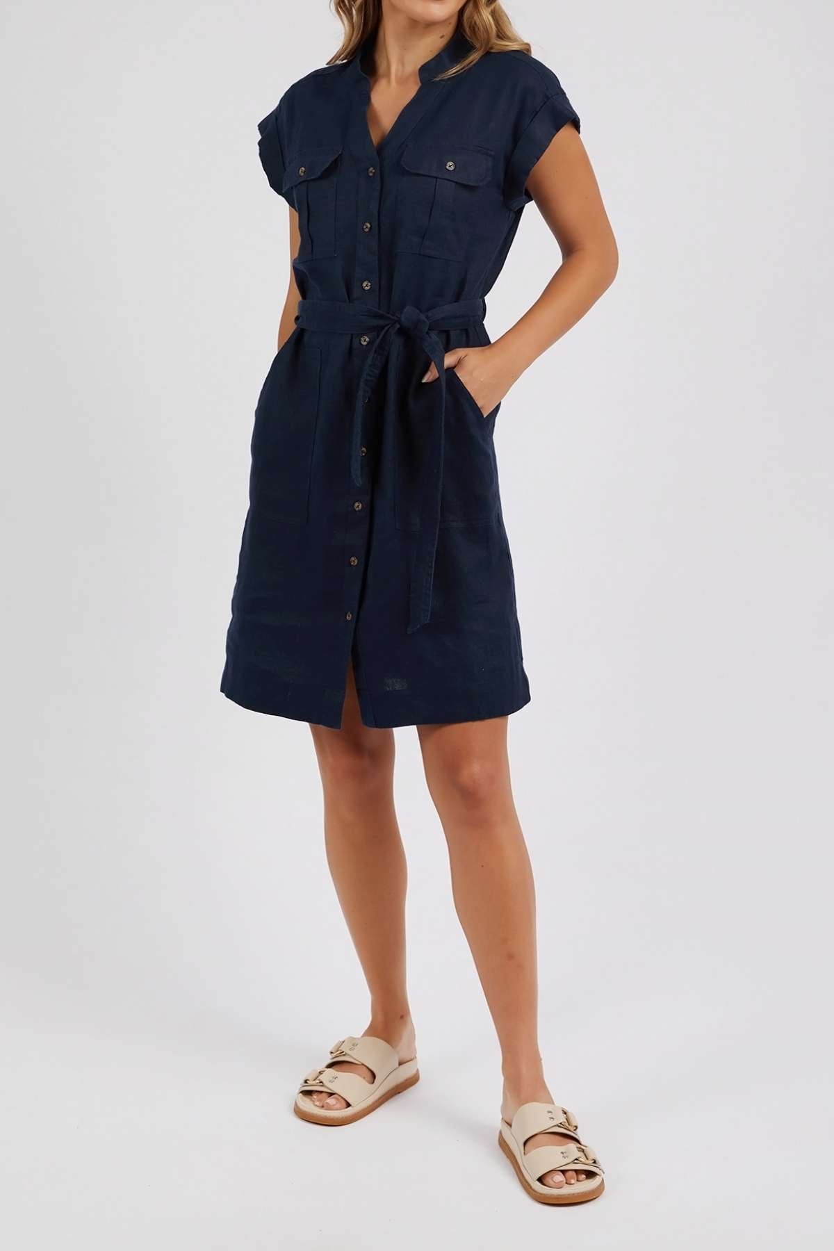 Woman in a short-sleeve, navy blue Foxwood Harlow Dress with v-neckline, waist tie, and pockets stands against a white background. Her look is finished with beige sandals featuring gold buckles.