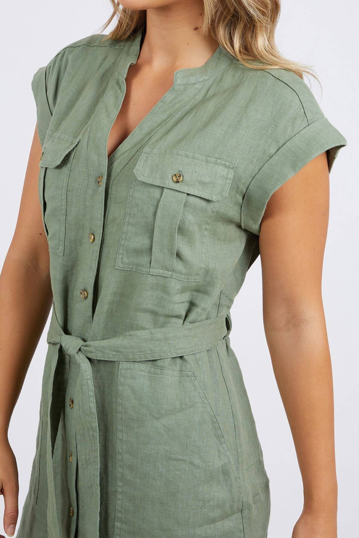 A woman wears the Foxwood Harlow Dress—a light green, short-sleeved, collarless linen shirt dress with front buttons, chest pockets, a belt, and a relaxed fit. Only her torso and part of her face are visible.