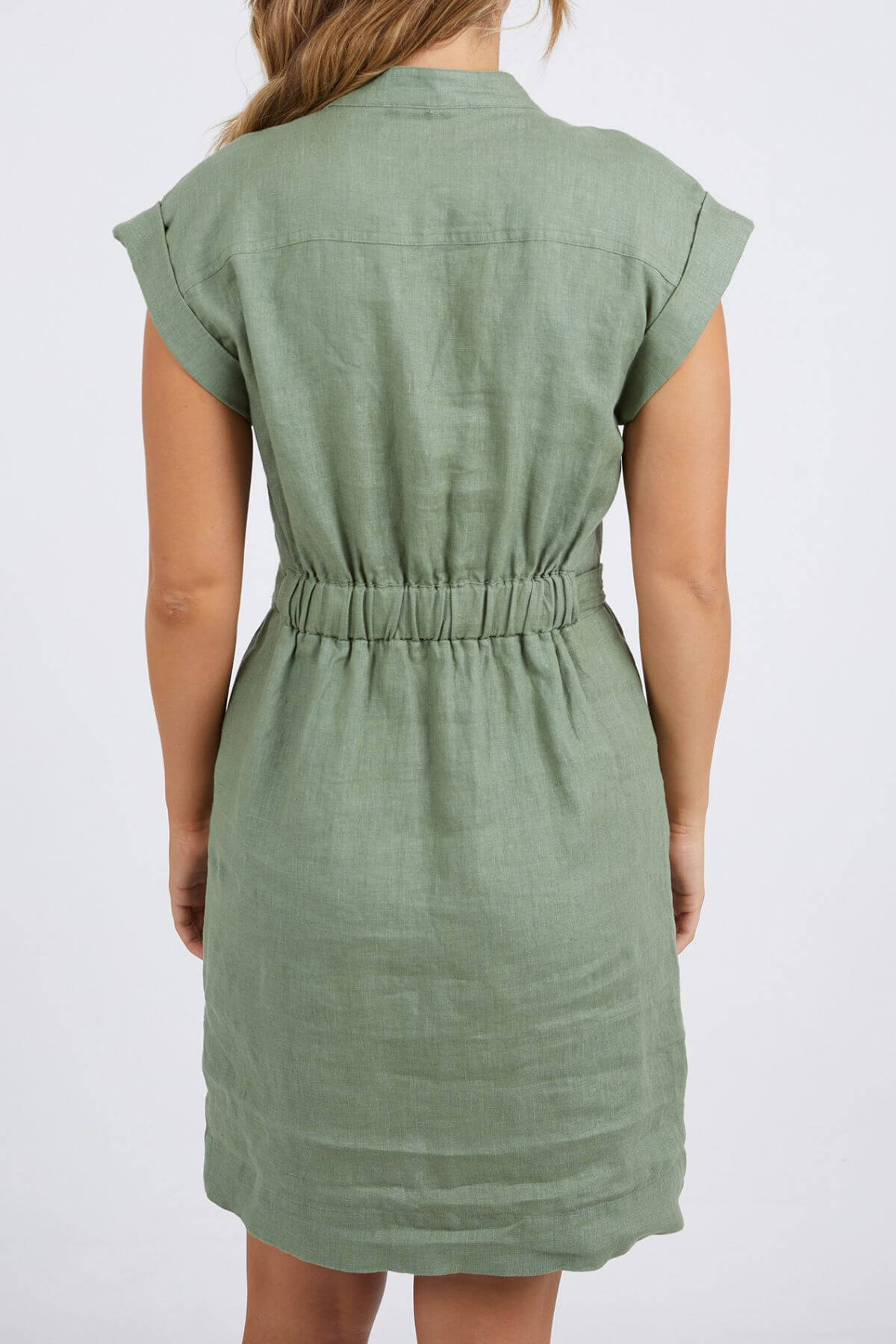 A person is shown from the back wearing the Foxwood Harlow Dress in light green. This linen summer dress has rolled sleeves, a straight collar, and an elastic waist. The background is plain white for a fresh, smart-casual look.