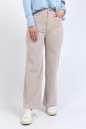 A person wears the Foxwood Henrietta Cord Pant in light beige, paired with a light blue long-sleeve shirt and white sneakers, standing against a plain white background with their hand in the pant’s front pocket.