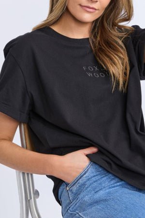 A woman sits on a chair wearing the Foxwood Huntleigh Oversized Tee in black with blue jeans. Her hair is loose and one hand is in her pocket. The cropped image shows her from shoulders to mid-thigh.