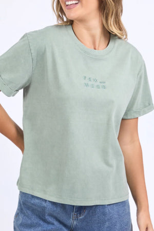 A smiling woman wears the Foxwood Huntleigh Oversized Tee in sage green with "FOX WOOD" embroidered on the front, styled with blue jeans.