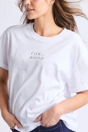 A woman with wavy hair models the Foxwood Huntleigh Oversized Tee in premium cotton, featuring small "FOX WOOD" text. She pairs it with dark blue jeans and stands with one hand in her pocket, highlighting the tee’s relaxed fit.