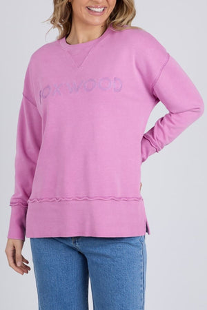 A woman smiles with one hand on her hip, wearing the Foxwood Illusion Crew—a relaxed light purple sweatshirt with the Foxwood logo embroidered on the front—paired with blue jeans.
