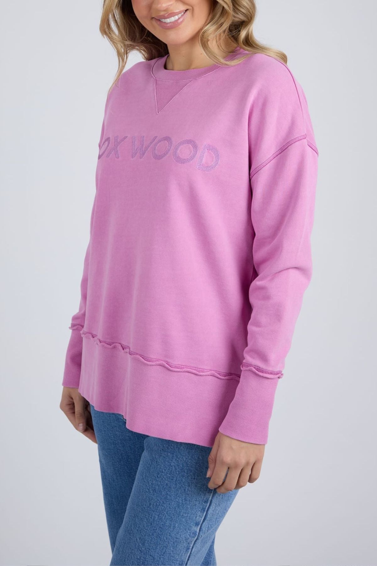 A woman wears the Foxwood Illusion Crew, a light pink relaxed sweatshirt made from soft cotton fleece with "OAKWOOD" text, paired with blue jeans. She stands against a plain light background, smiling slightly.