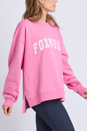 A person wearing a bright pink oversized Foxwood Interval Crew sweatshirt with "FOXWOOD" in large white letters, paired with dark leggings, stands against a plain light background. This relaxed-fitting crew is crafted from 100% cotton for ultimate comfort and style.