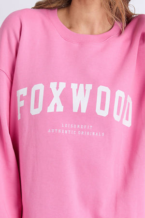 A person is wearing the Foxwood Interval Crew, a relaxed-fit pink crew sweatshirt made from 100% cotton Foxwood LeisureFit fleece. The sweatshirt features "FOXWOOD" in large white letters with "LEISUREFIT AUTHENTIC ORIGINALS" in smaller text underneath. Only the upper torso is visible.