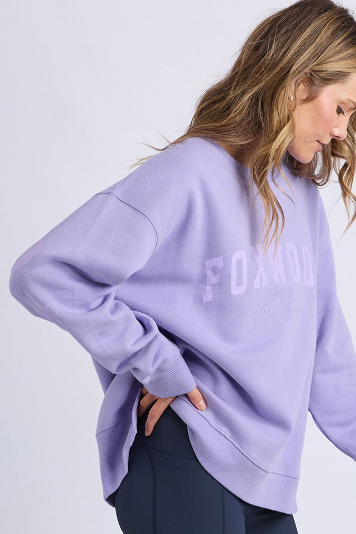 Wearing the Foxwood Interval Crew—a loose, lavender sweatshirt crafted from 100% cotton LeisureFit fleece—a woman stands sideways against a light background in dark leggings, smiling gently.