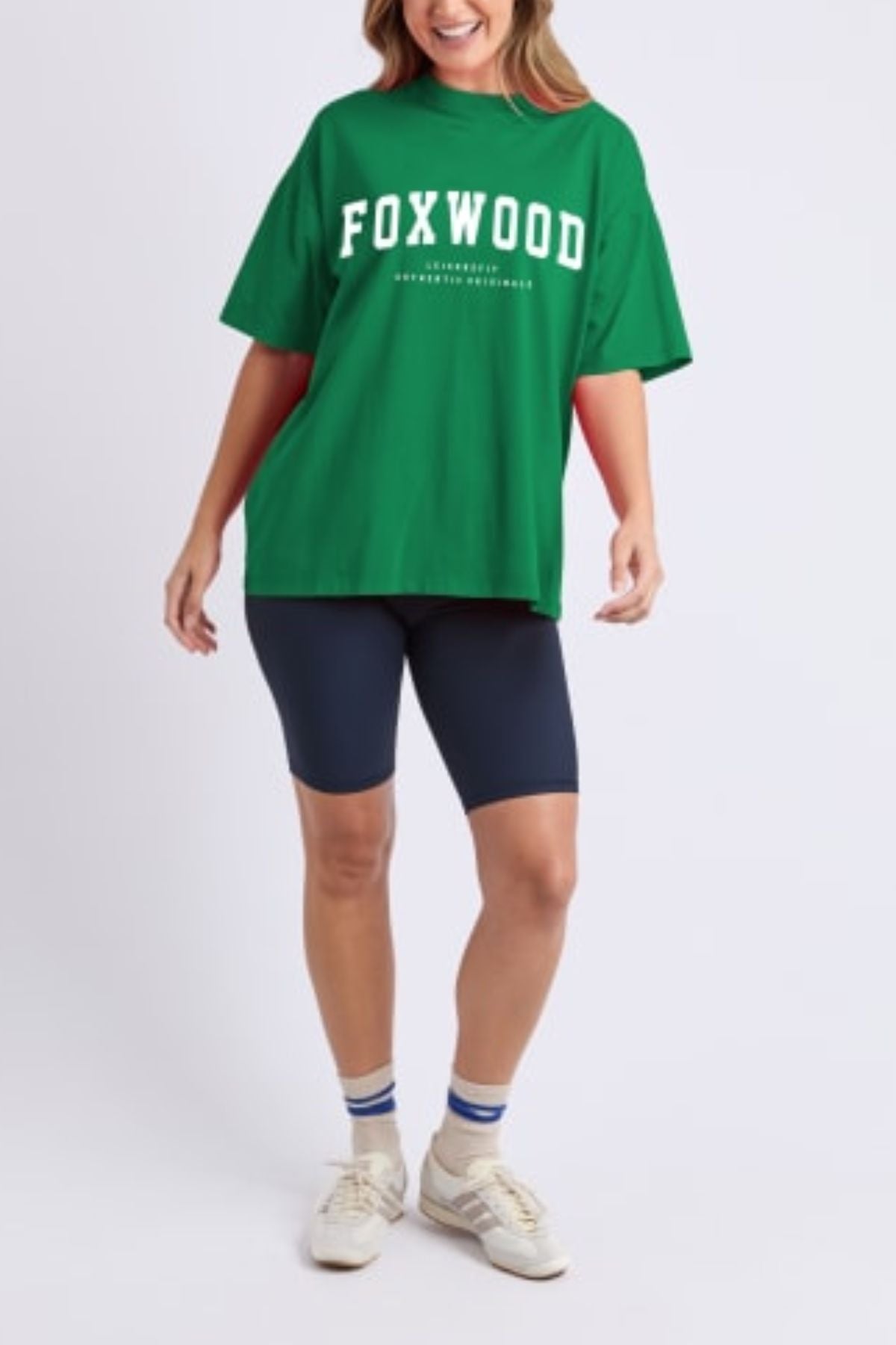 Wearing a loose green Foxwood Interval Tee, navy blue bike shorts, white striped socks, and white sneakers, a person smiles against a plain light background—showcasing effortless casual style.