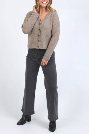 A woman smiles against a white background, touching her hair while wearing the relaxed Foxwood Jensen Button Cardi with tortoiseshell buttons, dark gray wide-leg jeans, and black heeled boots.