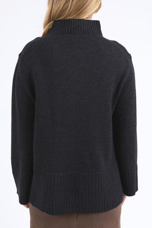 A person with long blonde hair, facing away from the camera, wears the Foxwood Jude Knit—a relaxed fit, dark ribbed mock neck sweater—paired with a brown skirt against a light background.