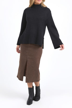 A person wearing the Foxwood Jude Knit, a relaxed fit black mock neck sweater, paired with a brown midi skirt with a front slit and black ankle boots, stands against a plain white background.