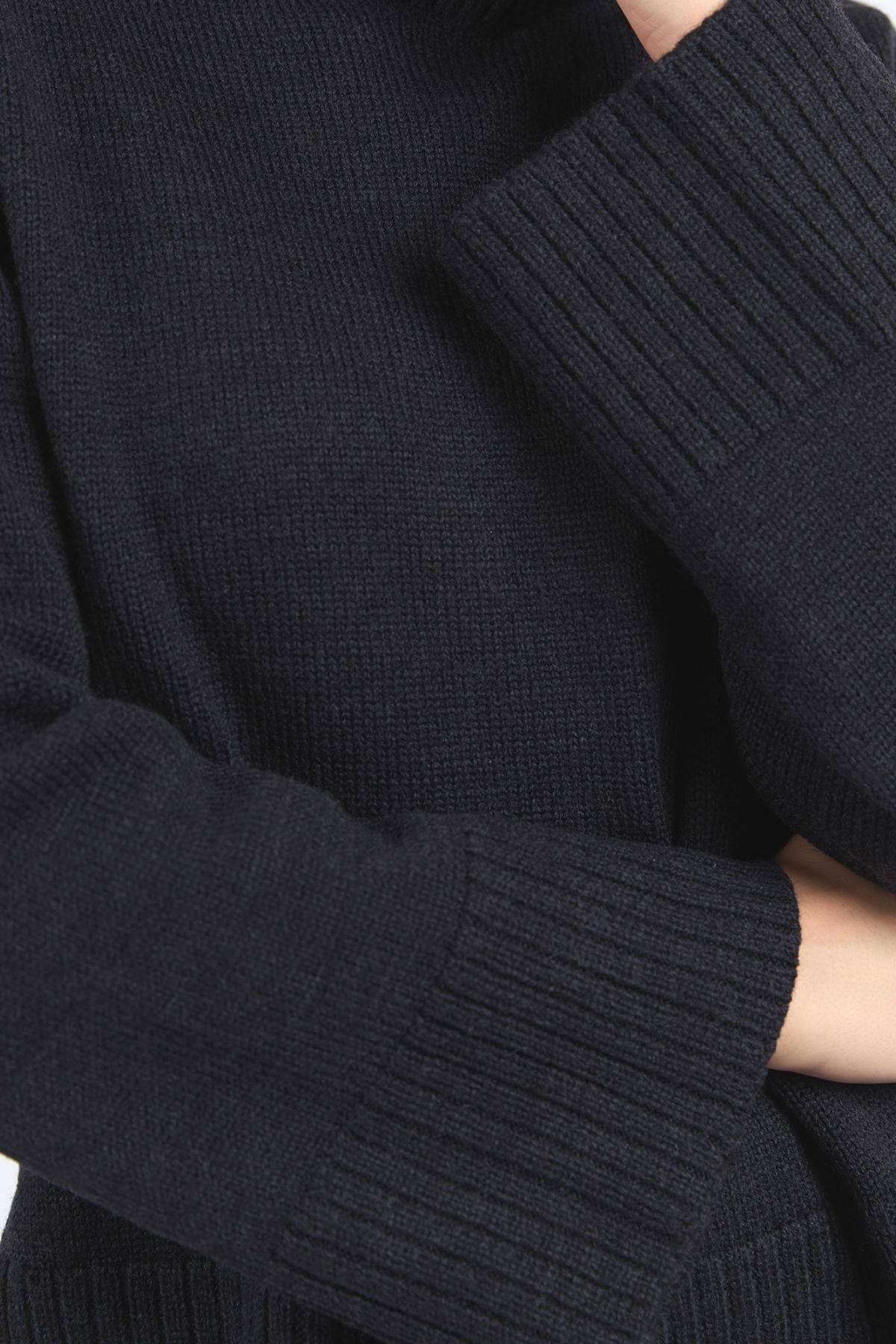 Close-up of a person wearing the Foxwood Jude Knit, a dark ribbed mock neck sweater with long sleeves and a relaxed fit, draping comfortably and showing only part of their body with crossed arms.