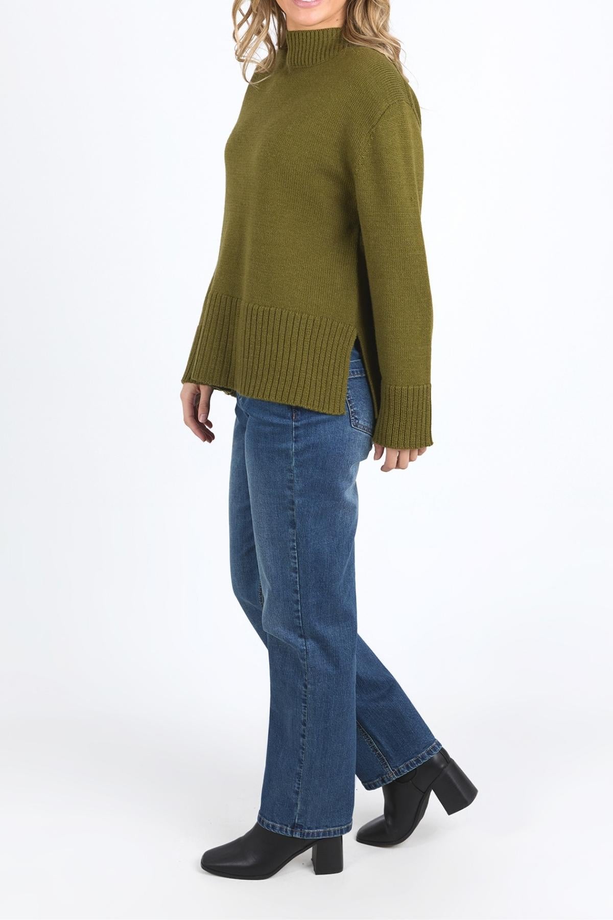 A person stands against a plain white background wearing the relaxed fit Foxwood Jude Knit in olive green with a mock neck, ribbed hem and sleeves, paired with blue jeans and black heeled ankle boots.