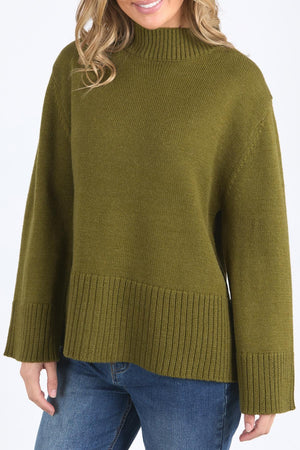 A woman models the Foxwood Jude Knit, a relaxed fit olive green sweater with a mock neck, paired with blue jeans. Only her torso and lower face are shown against a plain background.