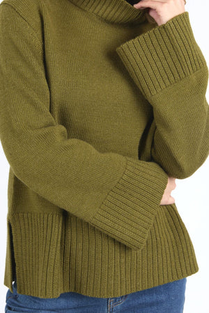 A person in a relaxed-fit, chunky olive green Foxwood Jude Knit sweater with wide ribbed cuffs and hem, paired with blue jeans. One hand touches their neck as their face is partly out of frame.