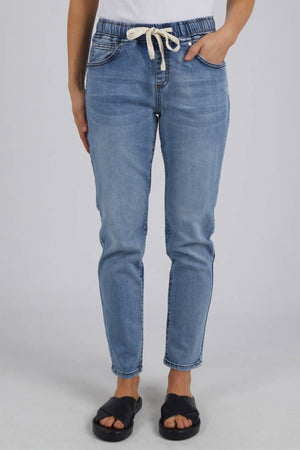 A person models the Foxwood Juliette Jogger Jean—light blue with an elastic waist—paired with a white top and black slide sandals, posed against a plain light background.