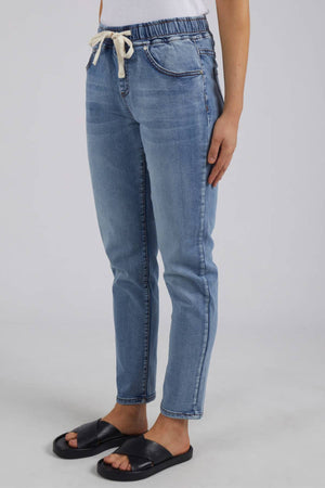 A person wearing Foxwood Juliette Jogger Jean in light blue, a white top, and black slide sandals stands against a plain light gray background.