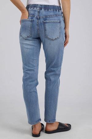 Someone stands facing away, wearing Foxwood Juliette Jogger Jeans—light blue with an elastic waist and two back pockets—with black slide sandals. The background is a plain light gray.