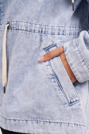A person wearing the Foxwood Kingston Anorak, a light blue, vintage washed Rigid Denim jacket with white drawstrings featuring metal tips, an adjustable waist cord, and their right hand partially in a front pocket.