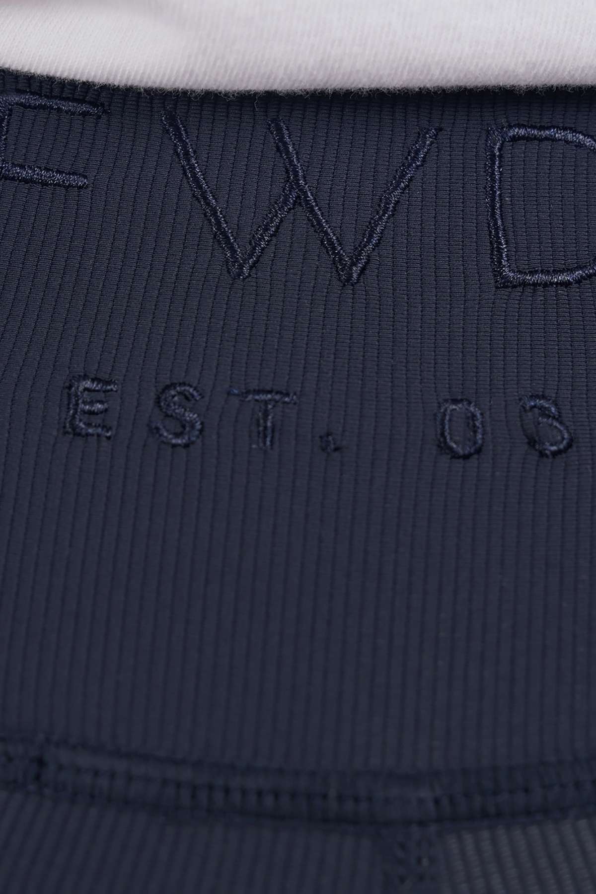 Close-up of dark ribbed fabric from the Foxwood Luxe Rib Short, featuring "EST. 03" embroidered text with part of larger letters above; elastic waist and a hint of white fabric at the top edge.