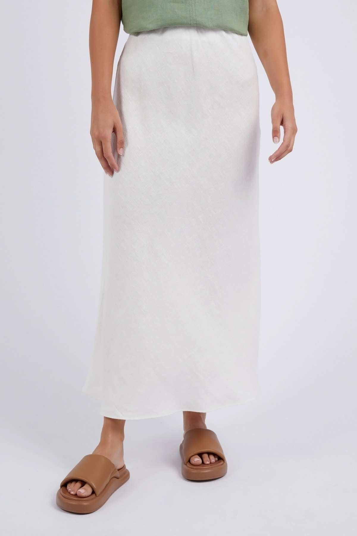 A person dons a light green top and a white Foxwood Lyon Slip Skirt with an elastic waist, standing against a plain background. They pair this ensemble with brown slide sandals, infusing casual elegance into their look.