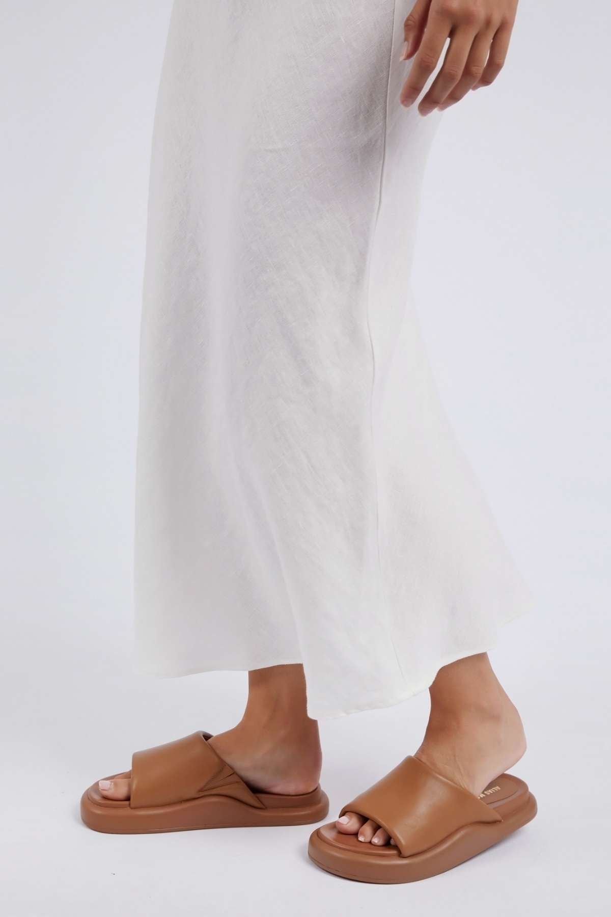 A person stands in a Foxwood Lyon Slip Skirt, highlighting their brown leather slide sandals with thick soles against a plain light background. The image showcases the lower part of the skirt and the stylish sandals.