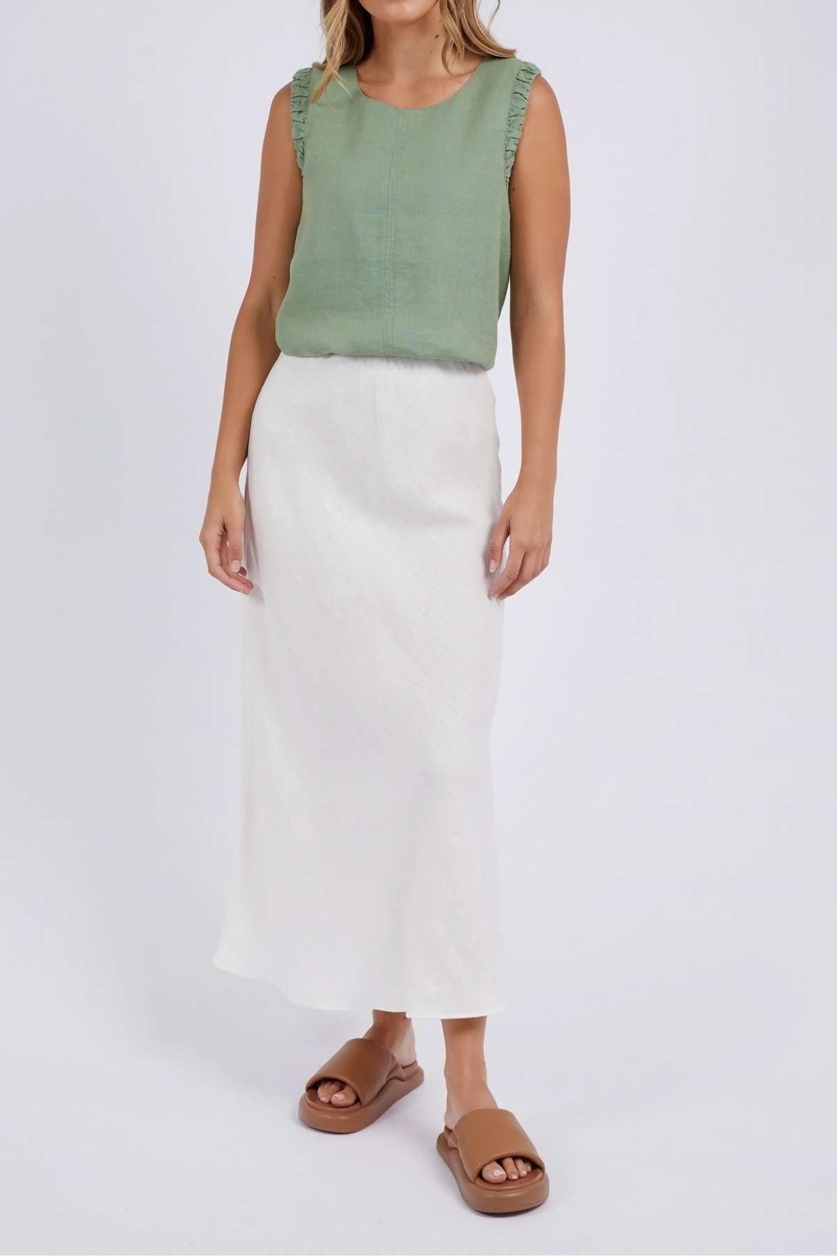 A person is in a sleeveless green top and the Foxwood Lyon Slip Skirt, which is a long white linen blend maxi skirt. They have long hair and are wearing tan sandals on a plain background.