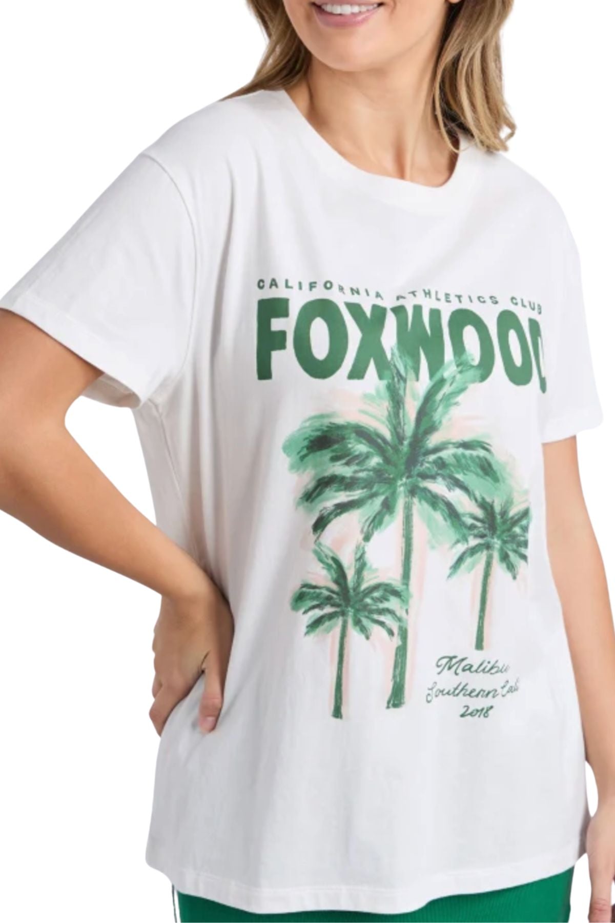 A woman in the Foxwood Malibu Tee, made from airy cotton, smiles with her hand on her hip. The white tee displays green palm trees and "FOXWOOD California Athletics Club Malibu Southern Cal 2017" on the front.