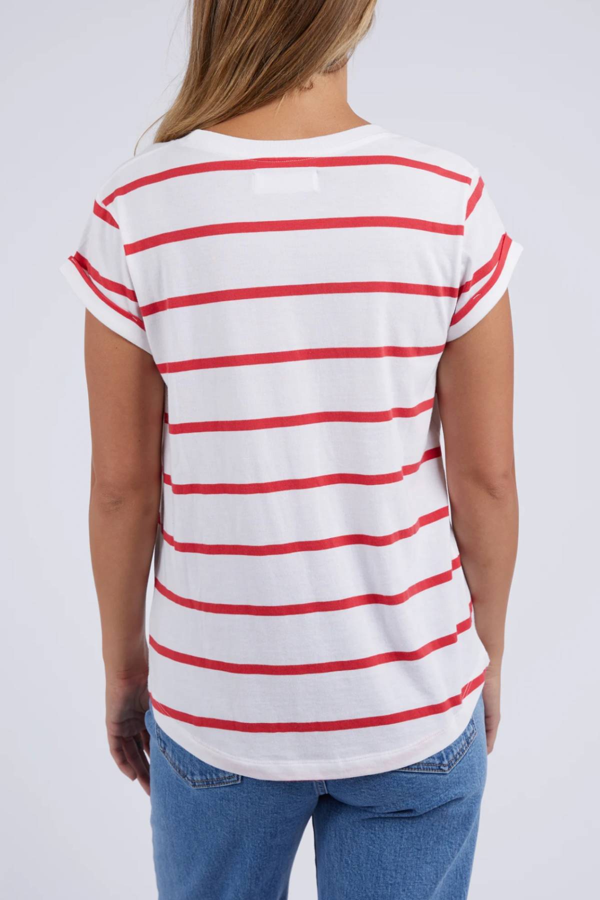 A fashion-forward individual with long hair is seen from the back wearing a Foxwood Manly Stripe Tee in soft Pima cotton jersey. The white T-shirt has red horizontal stripes and short, rolled-up sleeves, paired stylishly with blue jeans against a plain light gray background.
