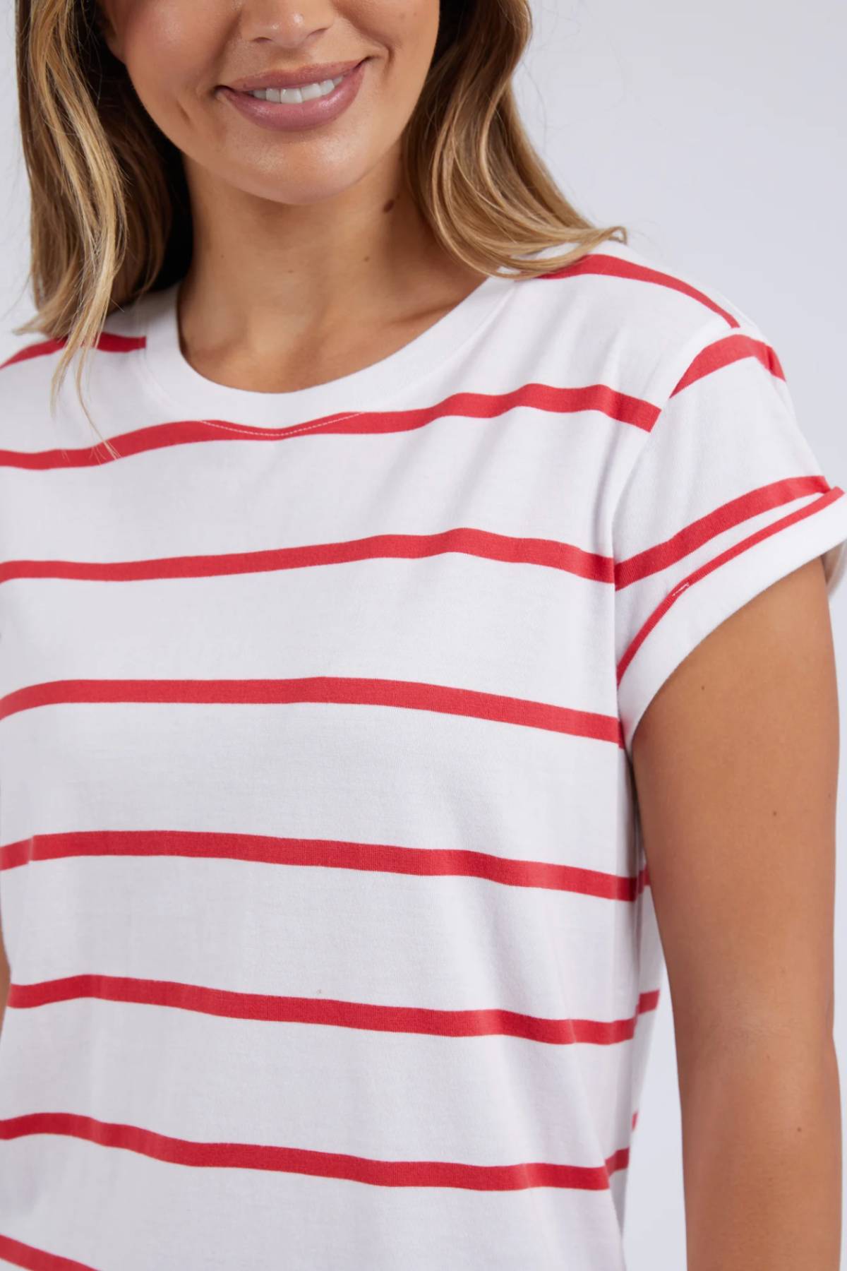 A person with long hair is wearing a stylish Foxwood Manly Stripe Tee made from Pima cotton jersey. This white shirt features red horizontal stripes and short rolled-up sleeves, creating a striking appearance against the plain, light-colored background.