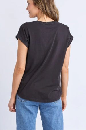 A person with light brown hair is shown from the back, wearing a Foxwood Manly Tee and light blue jeans, standing against a plain light background.