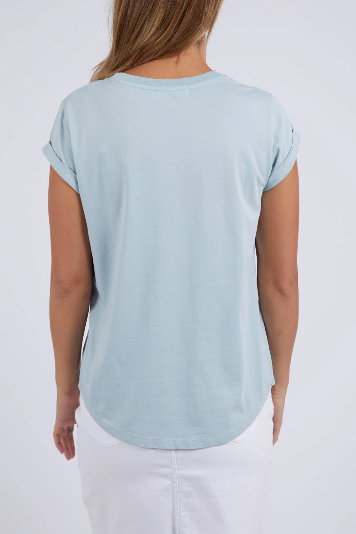 A woman stands with her back to the camera, wearing a light blue Foxwood Manly Tee and a white skirt against a plain light background for an easy, casual look.