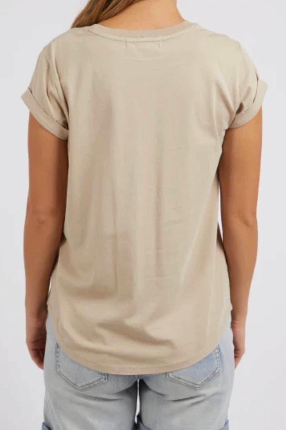 A person is seen from behind wearing the Foxwood Manly Tee—a beige, 100% cotton short-sleeve shirt with rolled cuffs—paired with light blue denim shorts against a plain light background for a casual chic look.