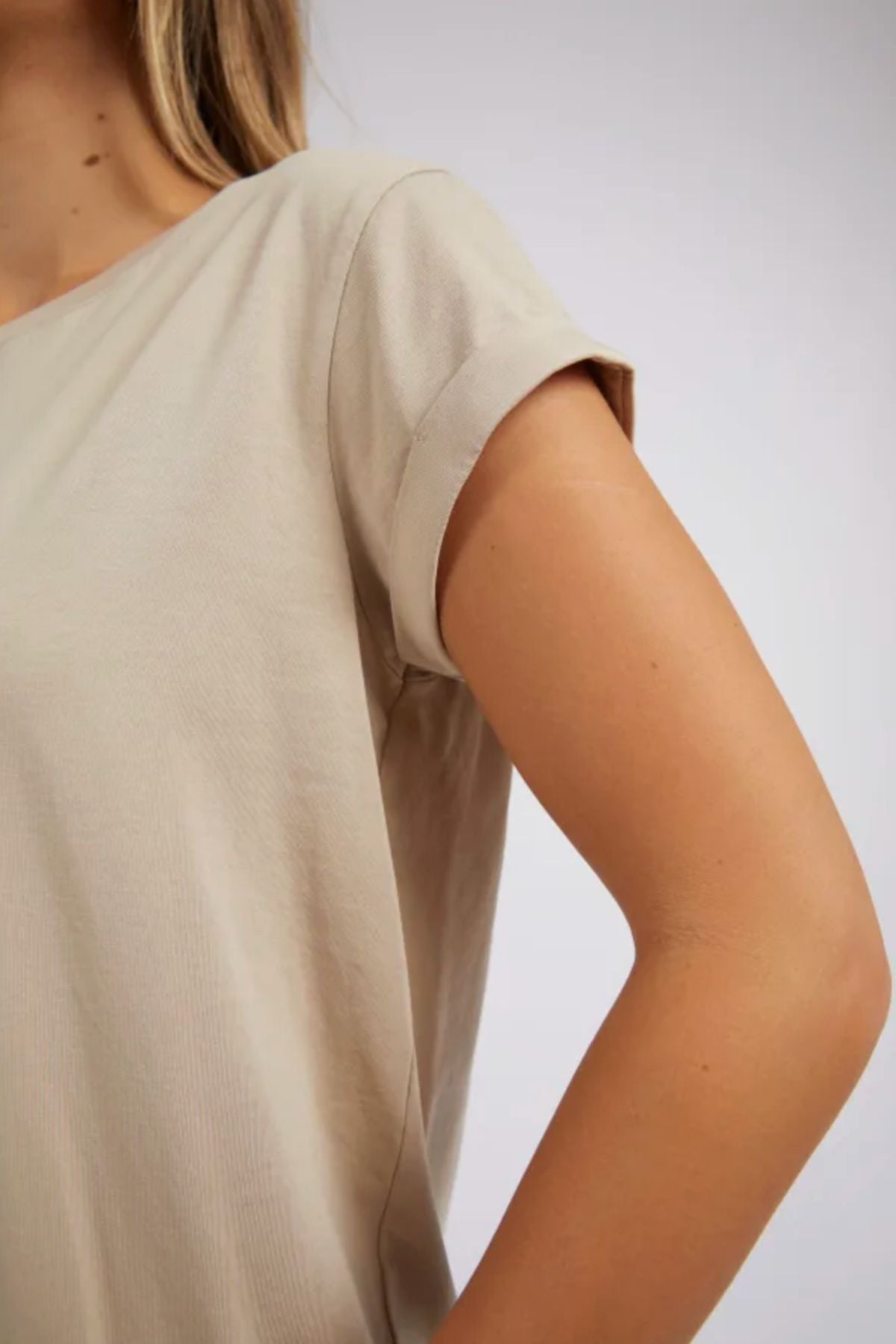 Model wears the Foxwood Manly Tee in beige, featuring short sleeves and a relaxed fit. With one arm bent and hand on hip, they showcase effortless casual style. Photo focuses on upper torso, arm, and partial face against a plain background.