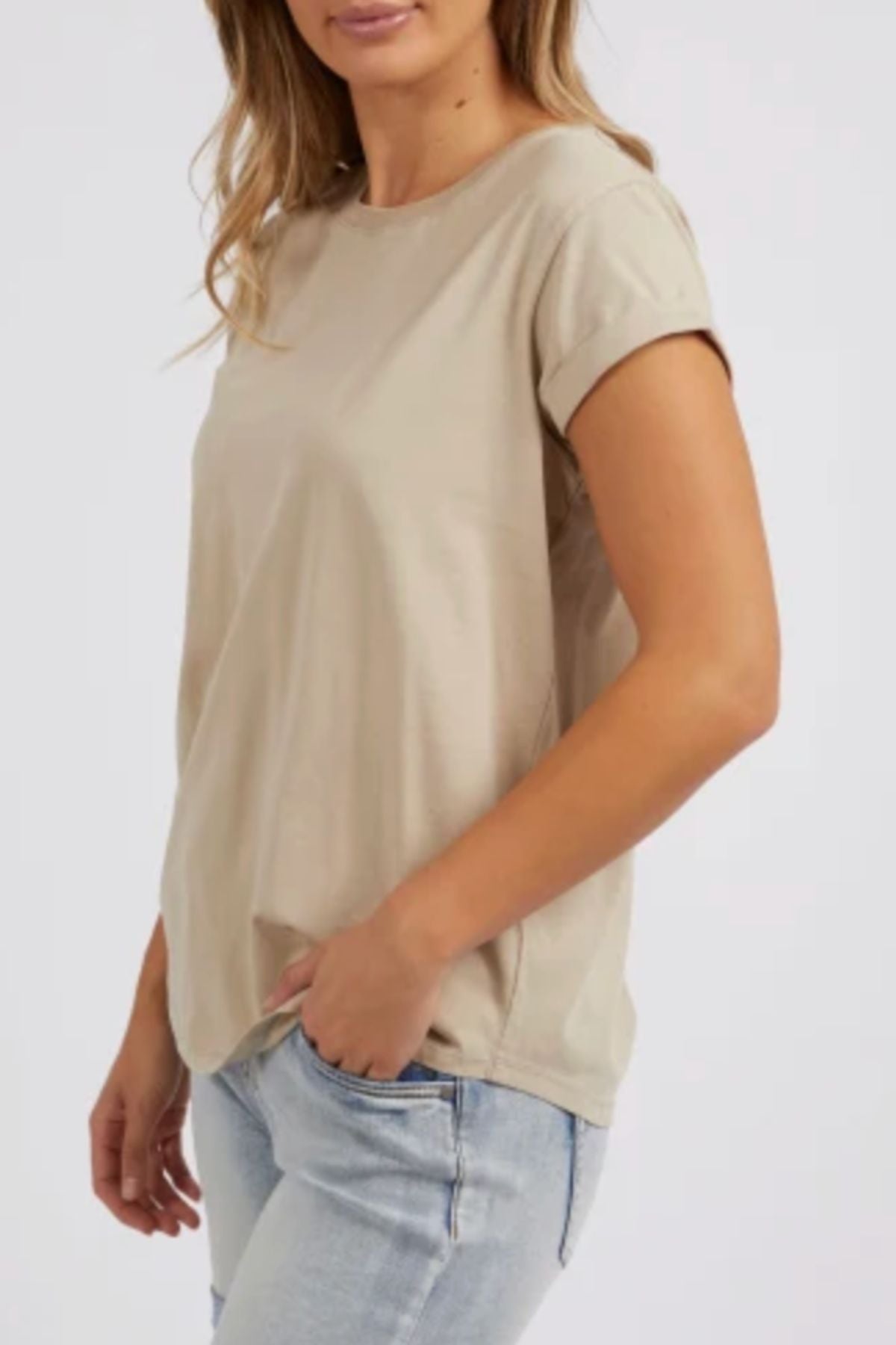 A woman poses with one hand in her jeans pocket, wearing the Foxwood Manly Tee in beige with light blue jeans. Her hair falls loosely as she stands against a plain, light-colored background.